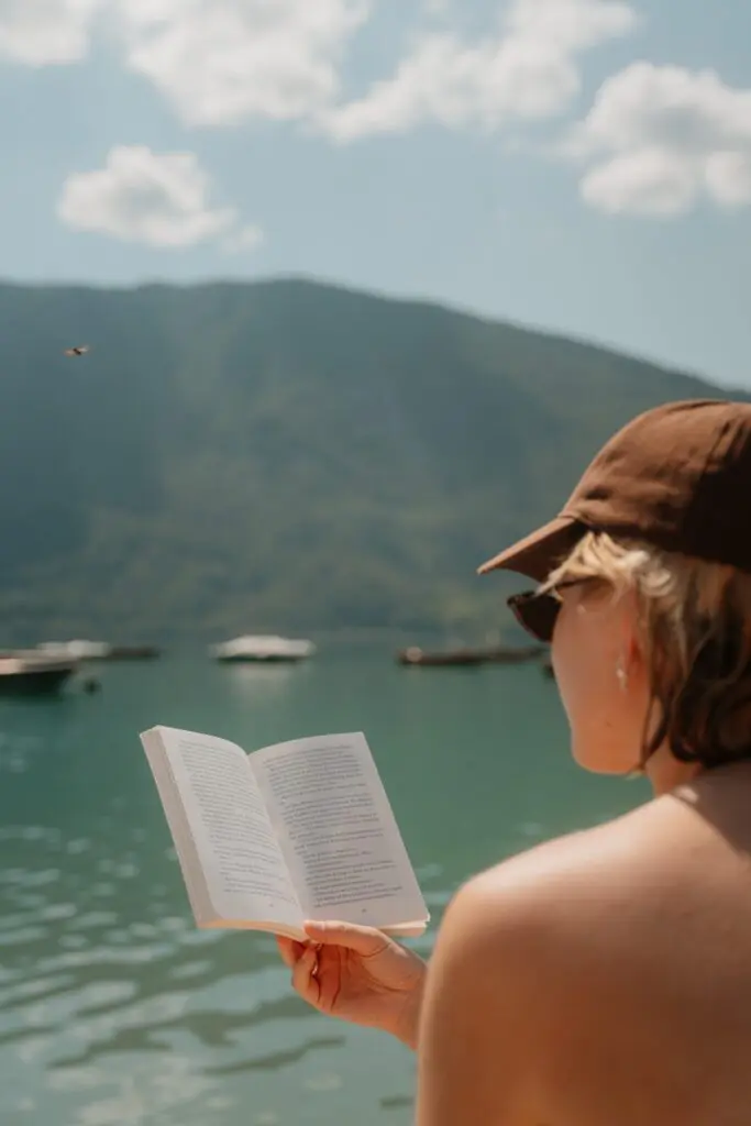 person reading a book by a lake with mountains