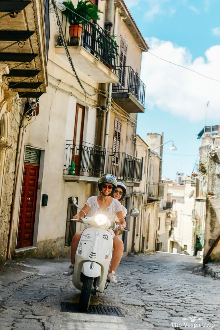 zwei Frauen auf einer weißen Vespa, die durch eine kopfsteingepflasterte Straße auf Sizilien fahren