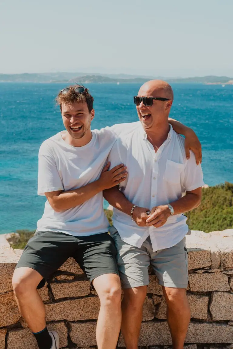 zwei Männer, die auf einer Steinmauer sitzen und mit Blick auf das Meer auf Sardinien lachen