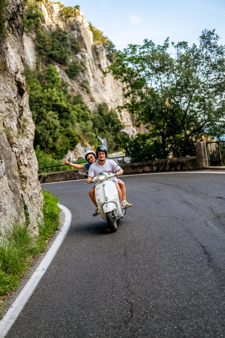 Zwei Personen fahren auf einer weißen Vespa entlang der Amalfiküste.