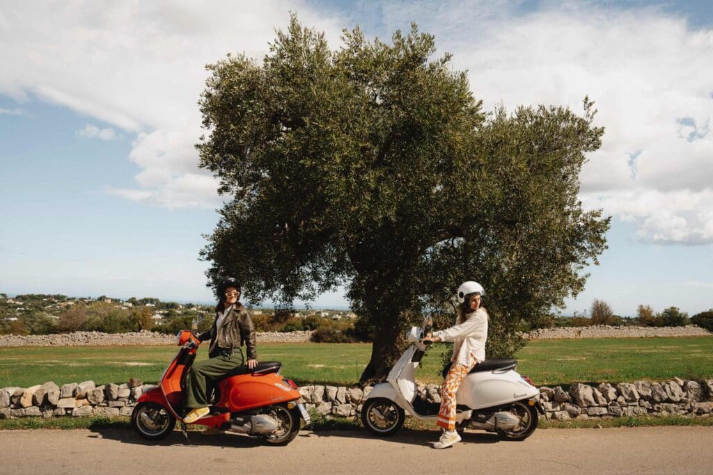 zwei Frauen auf Vespas vor einem Olivenbaum